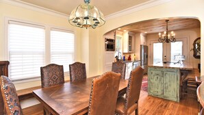 A dining area with a wooden table and chairs, a chandelier, and a kitchen in the background.