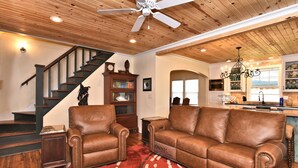 A spacious living room with a brown leather sofa, a wooden staircase, and a kitchen area with a chandelier.