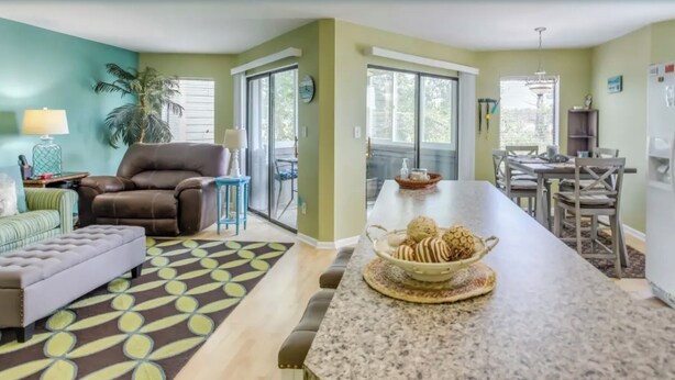 A modern kitchen with a central island, a dining area with a chandelier, and a living room with a sofa and a patterned rug. - North Myrtle Beach 2BR Condo – Pool, Fire Pit and Pet-Friendly (North Myrtle Beach)