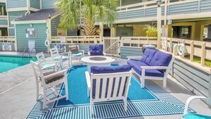A pool area with lounge chairs and a table, surrounded by a fence and palm trees. - North Myrtle Beach 2BR Condo – Pool, Fire Pit and Pet-Friendly (North Myrtle Beach)