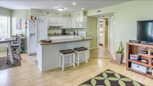 A modern kitchen with white cabinets, a central island with stools, a dining area with a table and chairs, and a living room with a TV and shelving. - North Myrtle Beach 2BR Condo – Pool, Fire Pit and Pet-Friendly (North Myrtle Beach)
