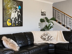 Ein schwarzes Ledersofa mit einer flauschigen Decke und einem Kissen, ein großes abstraktes Gemälde, ein Topfpflanze und eine Treppe mit Metallgeländer.