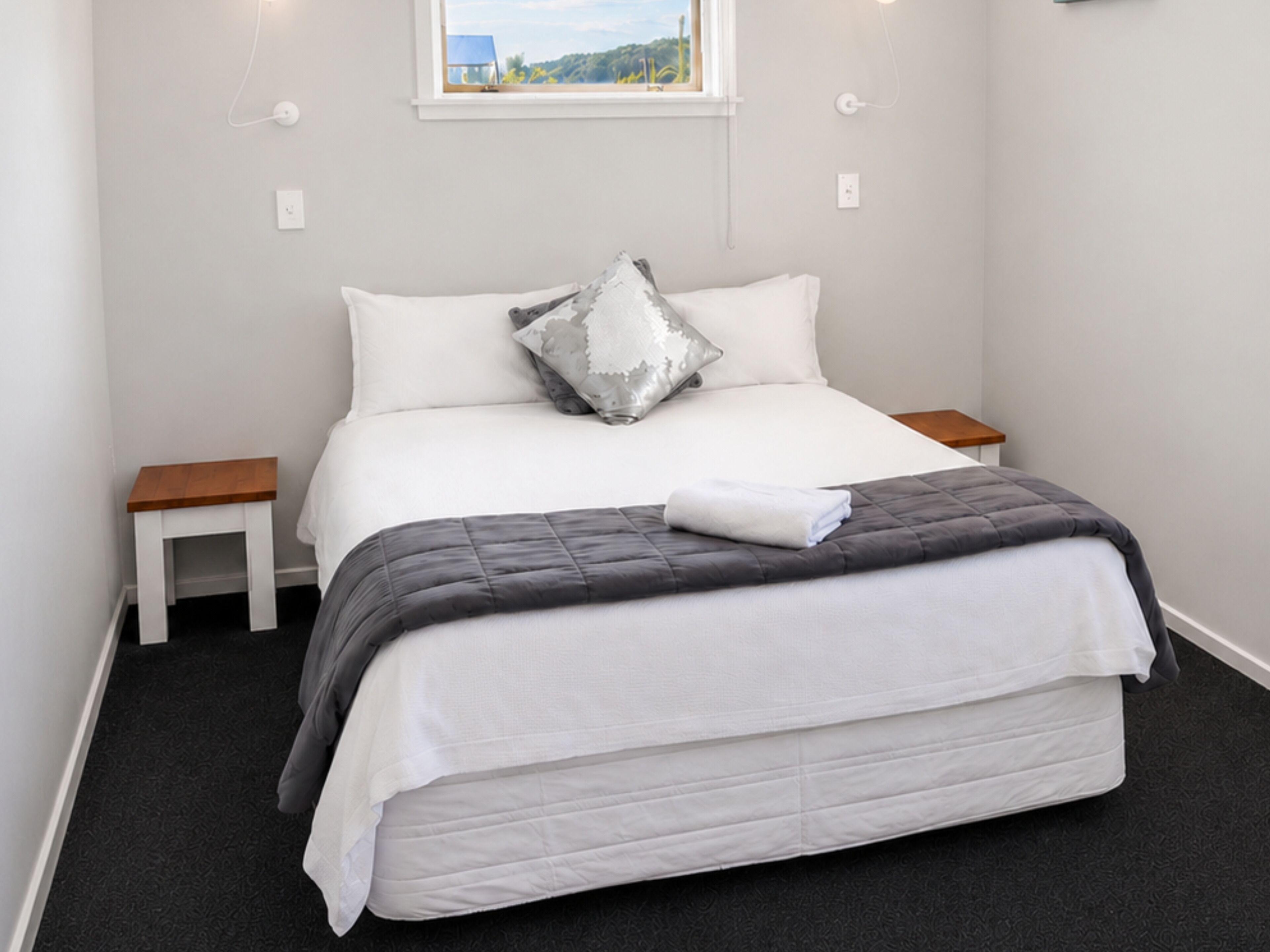 Un lit bien fait, recouvert d’une couette grise, avec des oreillers blancs et un coussin décoratif argenté. Il y a deux tables de chevet, une de chaque côté du lit.