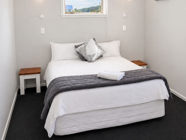 Un lit bien fait, recouvert d’une couette grise, avec des oreillers blancs et un coussin décoratif argenté. Il y a deux tables de chevet, une de chaque côté du lit.