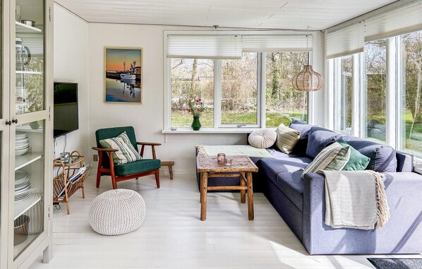 A sunlit room with a sofa, armchair, and round ottoman, a television, and a painting.