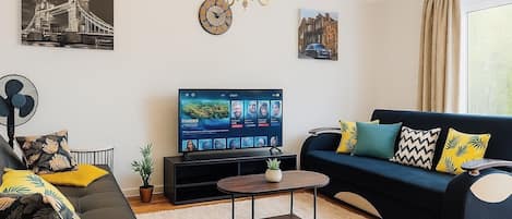 A modern living room with a black sofa, a coffee table, and a TV on a stand. There are decorative pillows and a plant on the sofa.