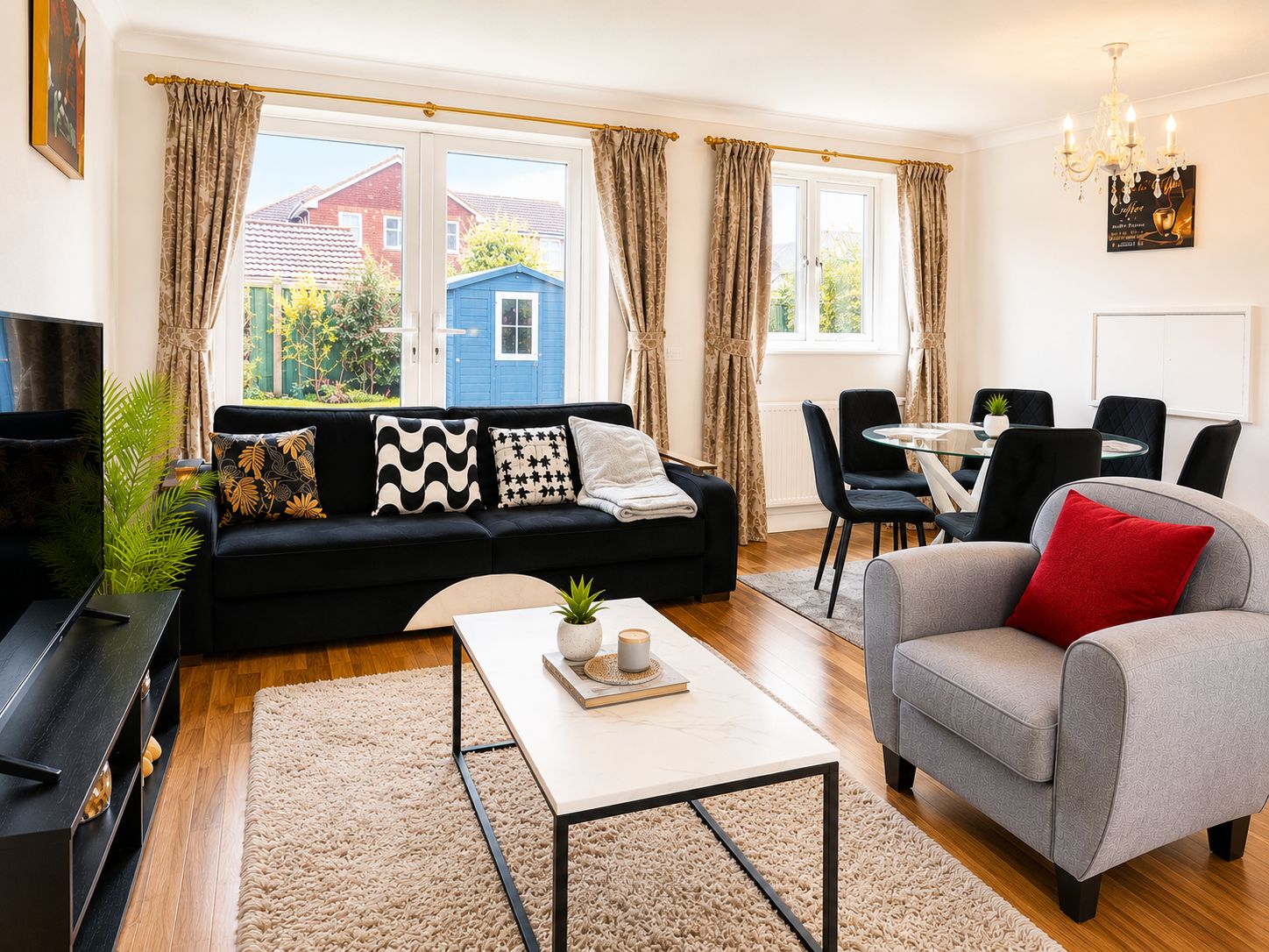 Een moderne woonkamer met een zwarte bank, een grijze fauteuil en een salontafel. Er is een eethoek met een tafel en stoelen, en een televisie op een dressoir.