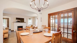 A dining room with a wooden table, chairs, and a chandelier. There is a fireplace and a view through a glass door. - Hot tub, 4bd Family Home - Bertrand Cottage (Manhattan)