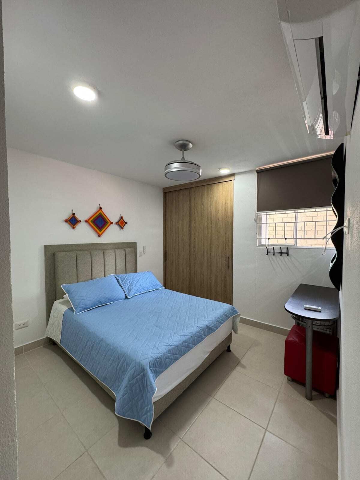 Un dormitorio con cama, armario, una mesita y ventana con persianas.