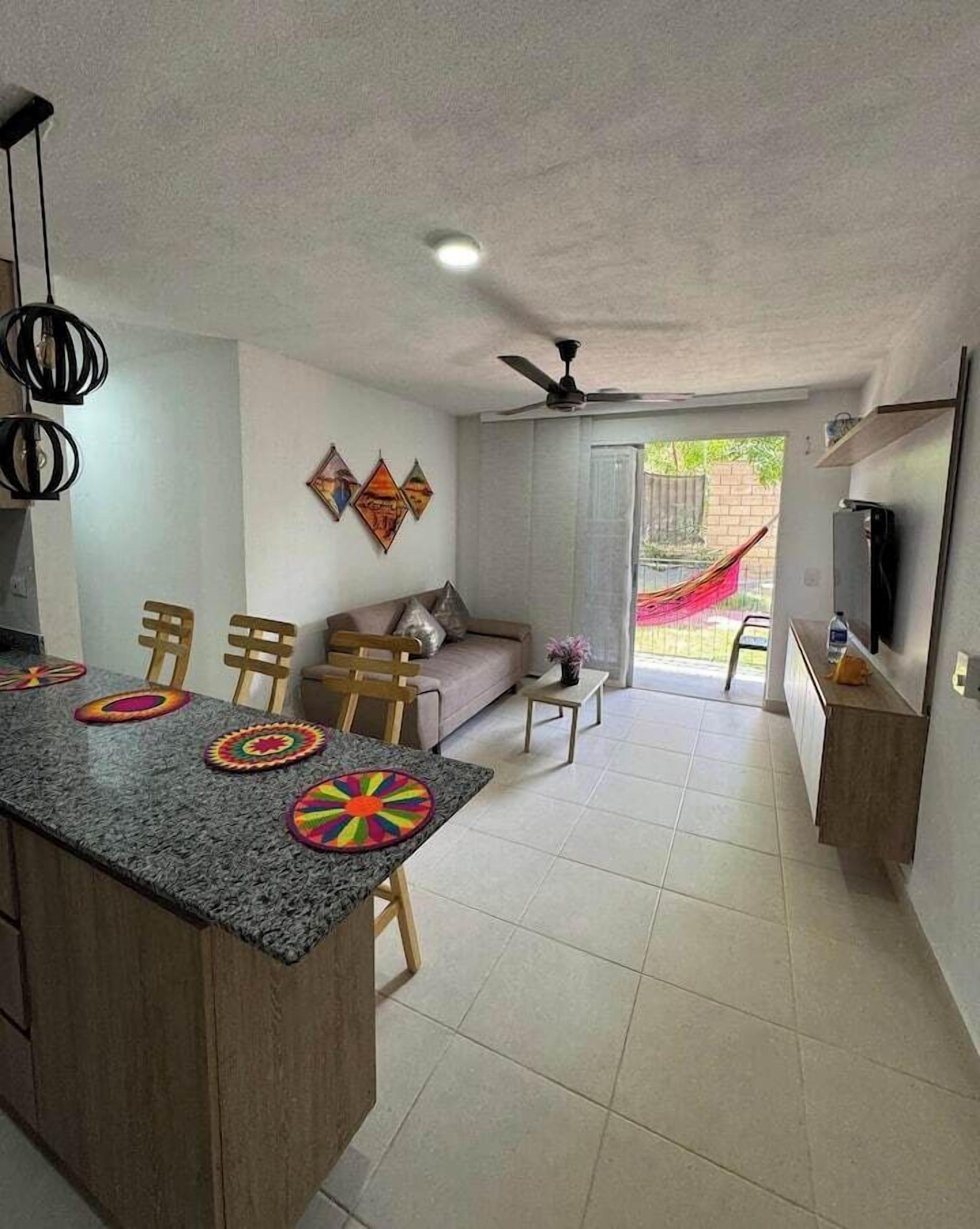 Une cuisine moderne avec un plan de travail en granit et des chaises en bois, un coin repas avec un canapé et un téléviseur, et une vue sur un jardin grâce à une porte vitrée.