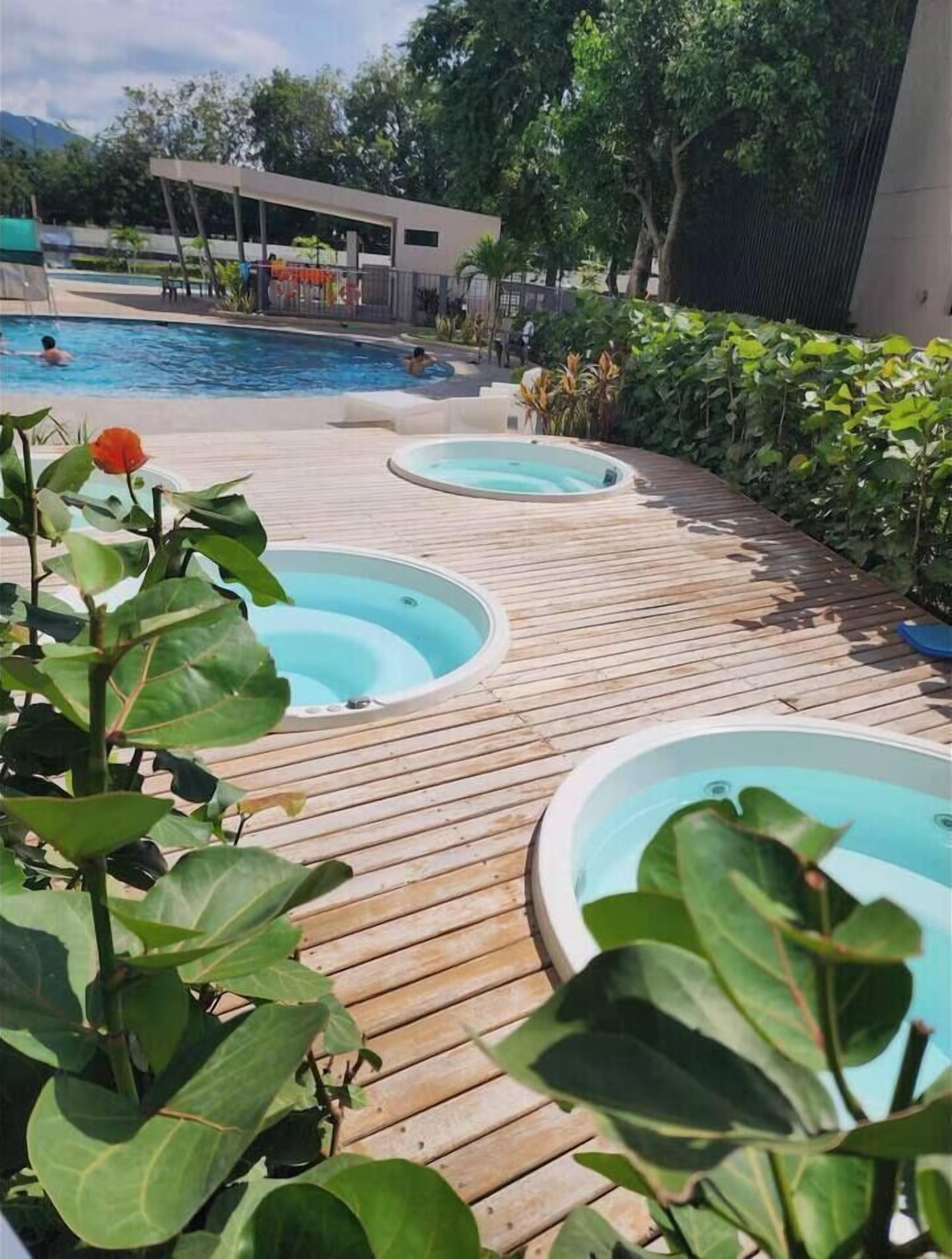 A wooden deck with three hot tubs, a swimming pool, and a modern building in the background.