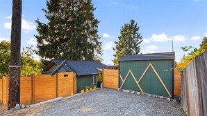 Ein Kiesweg führt zu einem Holztor, ein kleines Nebengebäude mit geneigtem Dach sowie ein größeres, grünes Gartenhaus mit Zickzackmuster.