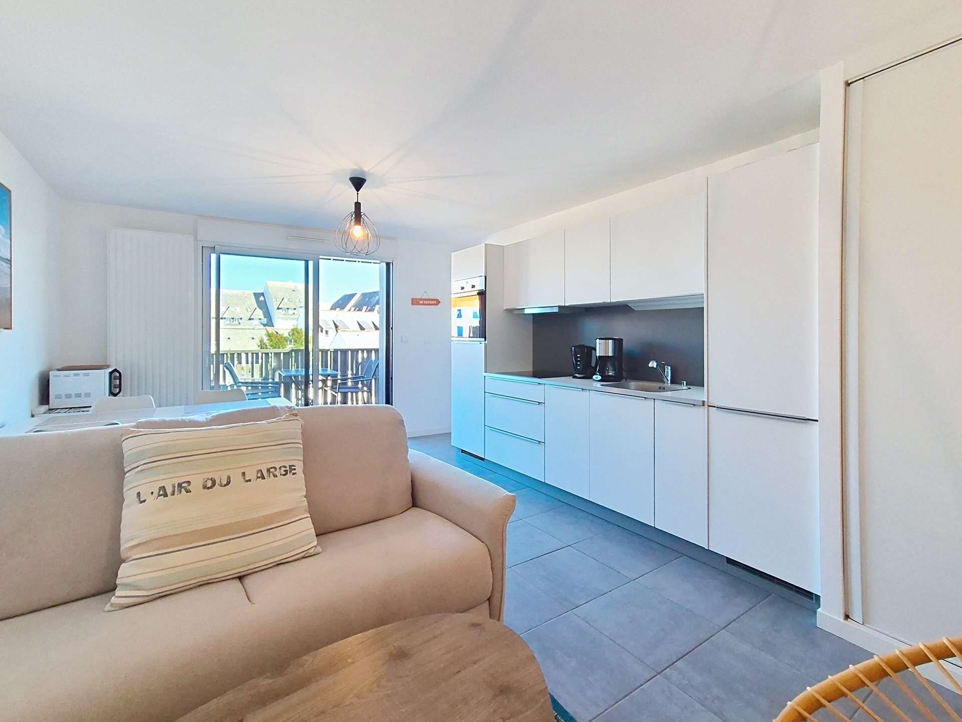 A modern apartment with a kitchenette, a sofa with a pillow, and a view of buildings through large windows.