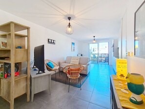 Un salon moderne avec un téléviseur, des fauteuils en osier, un canapé et un coin repas avec une table jaune.