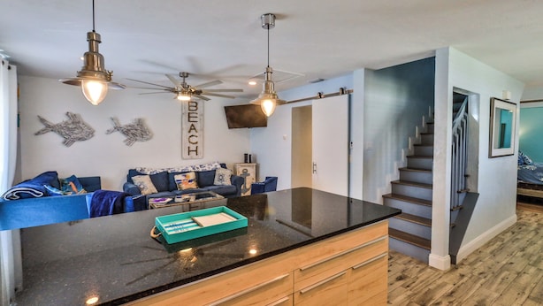 A kitchen with a black countertop, wooden cabinets, and a ceiling fan. There is a staircase leading to another level. - FLIP FLOP LIING 2! 4 Bdrms, 3 Bths, STEPS TO THE SAND! (New Smyrna Beach)