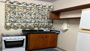 A compact kitchen with a white stove, wooden cabinets, a patterned curtain, and a small rug. - Tangará Inn in Ubatuba (Kitnet 1) (Taquaral Ubatuba)