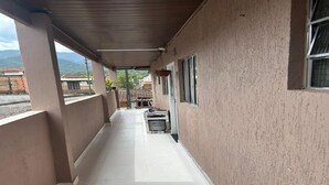 A covered corridor with a tiled floor, a window on the right, and a view of buildings and mountains in the distance. - Tangará Inn in Ubatuba (Kitnet 1) (Taquaral Ubatuba)