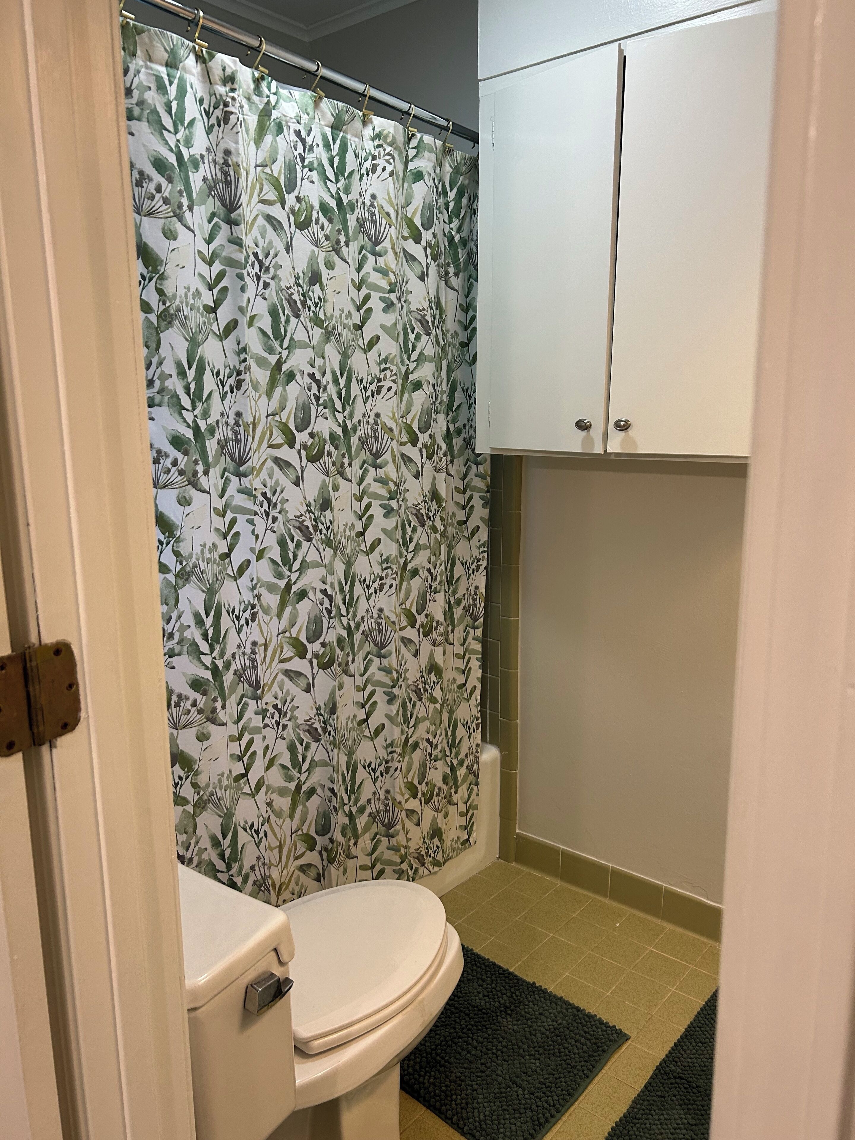 Ein Badezimmer mit Toilette, einer Duschvorhang mit floralem Muster und einem Schrank über der Toilette.