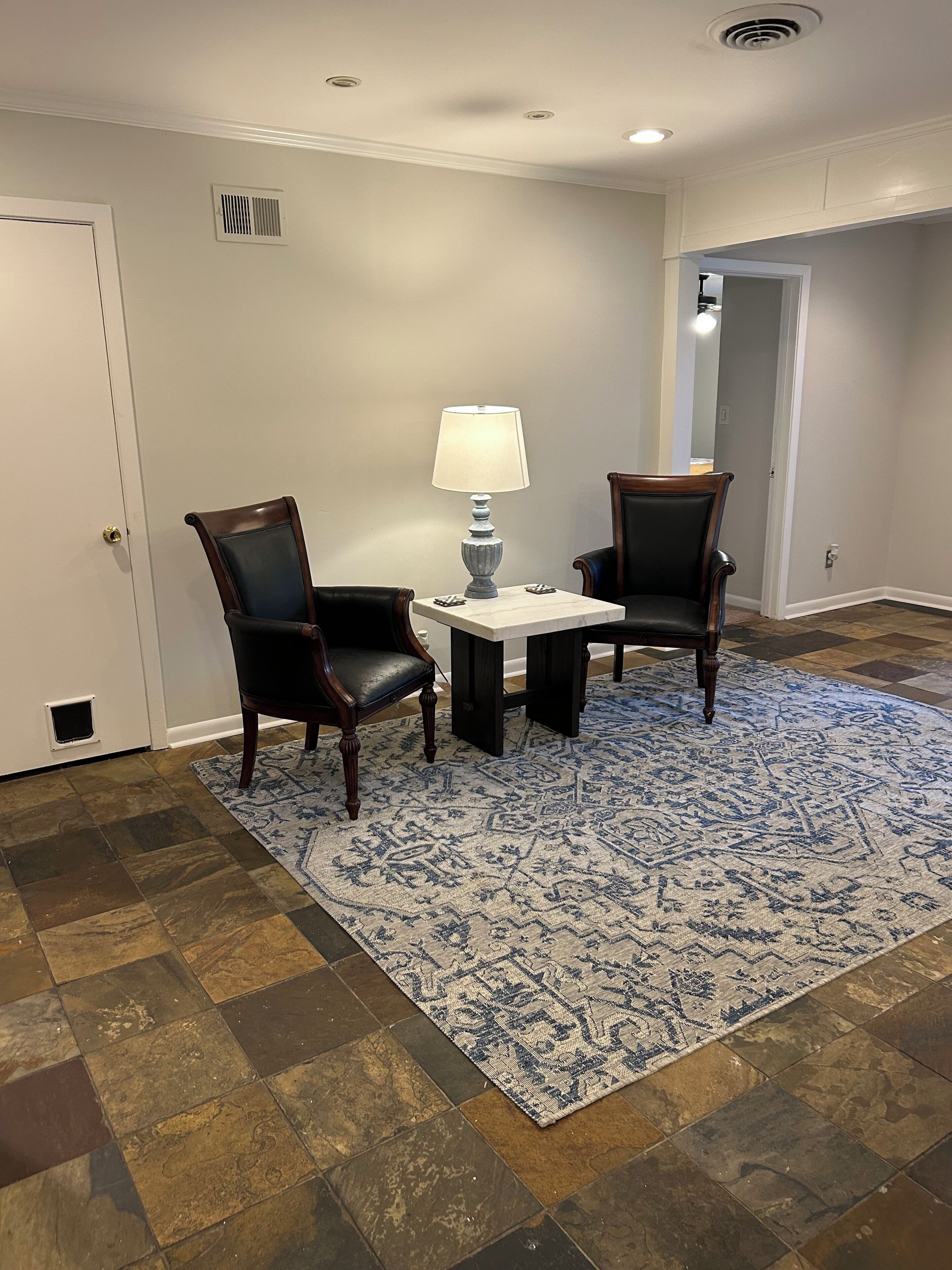 Une pièce avec un parquet en bois foncé, un tapis à motifs bleus, deux chaises en bois foncé, une petite table blanche avec une lampe et une porte blanche.