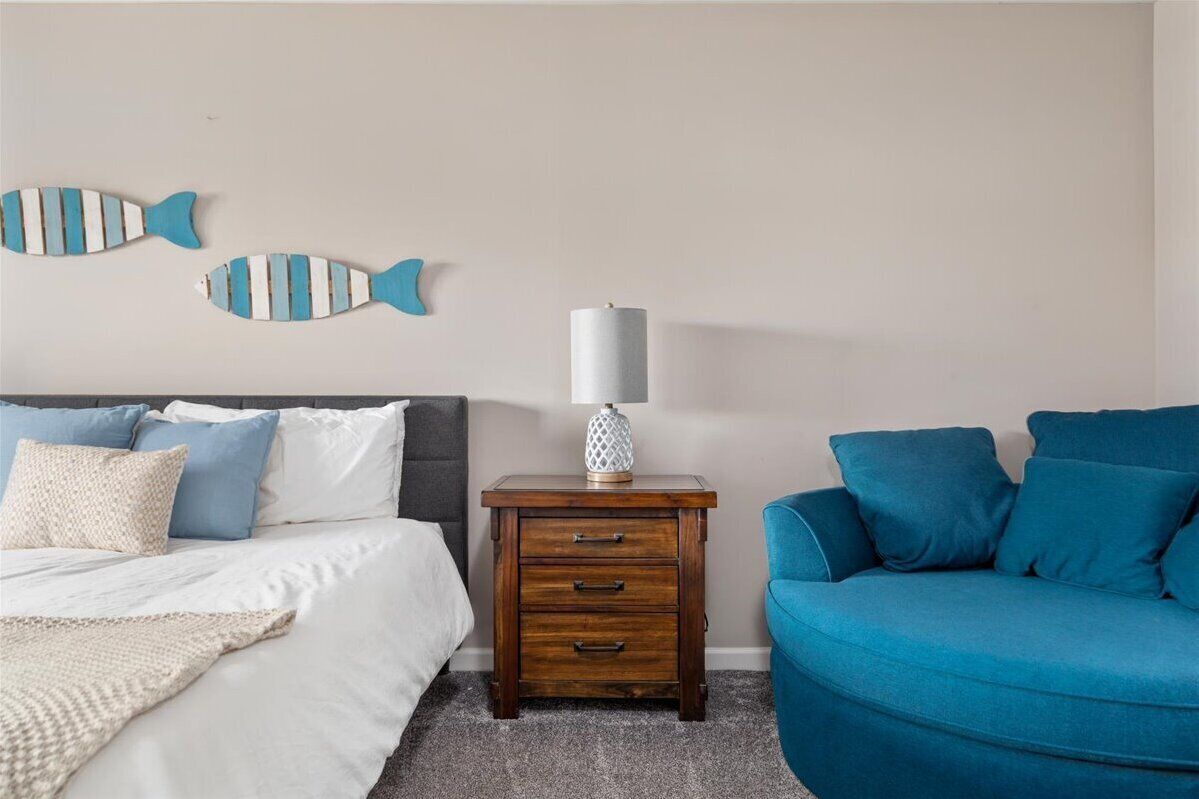 A bedroom with a bed, a nightstand with a lamp, a blue sofa, and two wall decorations.