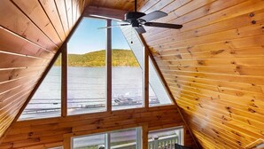 Techo de madera triangular con un ventilador y un ventanal grande que da a una masa de agua y colinas.