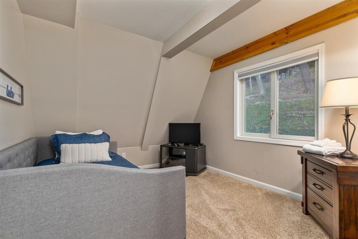 A bedroom with a sloped ceiling, a bed with a blue and white pillow, a TV stand, a wooden dresser, and a window with a view of trees.