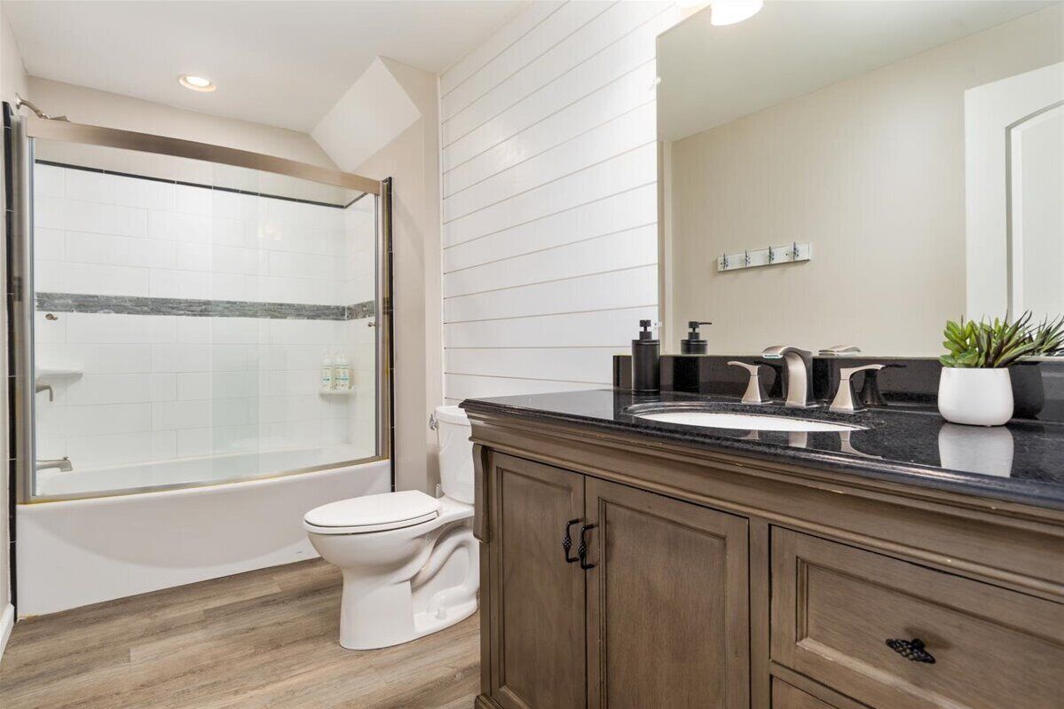 A modern bathroom with a glass-enclosed shower, a white toilet, a wooden vanity with a black countertop, and a large mirror.
