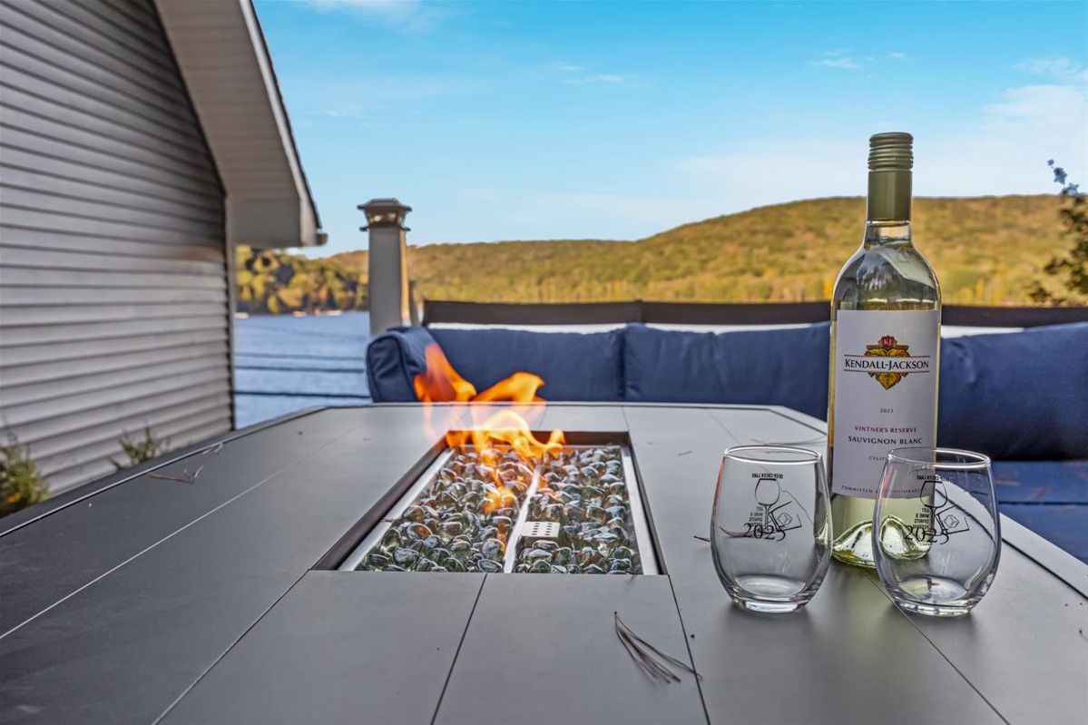A table with a fire feature, a bottle of wine, and glasses on a patio with a view of hills and water.