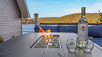 A table with a fire feature, a bottle of wine, and glasses on a patio with a view of hills and water.