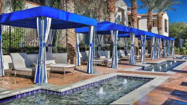 A pool area with multiple lounge chairs and blue canopies, surrounded by palm trees and a clear sky.