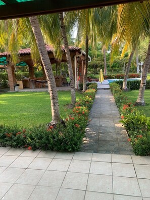 Un camí delimitat per palmeres i plantes florides, que condueix a una zona coberta amb seients i una piscina al fons.
