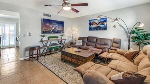 A living room with a brown leather sofa, a wooden coffee table, and a ceiling fan. There are two paintings on the wall, a plant, and a staircase leading to another room. - Havasu War Eagle - Pool, Games, Serene Stay (Lake Havasu City)