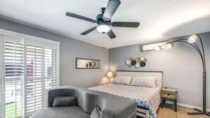 A bedroom with a bed, a gray armchair, a ceiling fan, a bedside table with a lamp, and a wall-mounted light fixture. - Havasu War Eagle - Pool, Games, Serene Stay (Lake Havasu City)