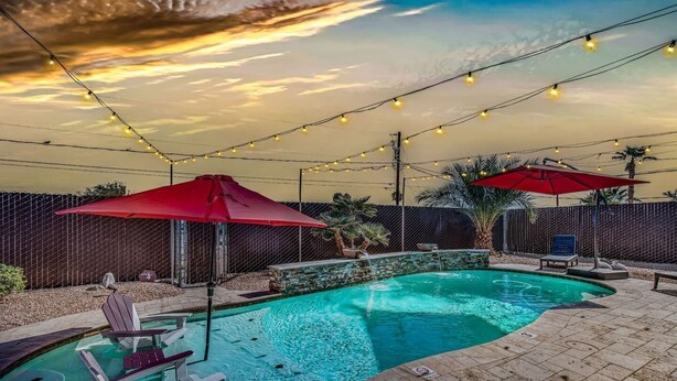 A swimming pool with a stone edge, surrounded by a brick patio. There are lounge chairs and umbrellas, and string lights overhead. - Havasu War Eagle - Pool, Games, Serene Stay (Lake Havasu City)