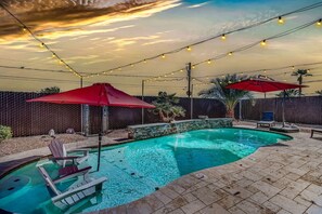 Una piscina con borde de piedra, rodeada por un patio de ladrillo. Hay sillas de descanso y sombrillas, además de luces colgantes.