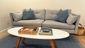 Un salón con un sofá gris, dos almohadas estampadas, una bandeja de mimbre con un control remoto, un libro y una lupa sobre una mesa de centro blanca con patas de madera.