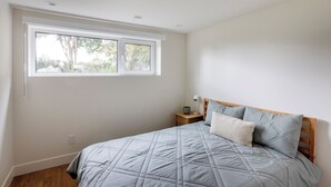Un dormitorio con un ventanal grande, una cama con colcha acolchada, una lámpara de noche y una mesita de noche.