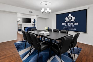 A conference room with a projector, black chairs, a blue carpet, and a large screen displaying 'The Flowers Retreat' logo.