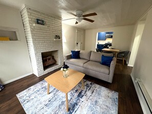 A living room with a fireplace, a sofa, a coffee table, and a kitchen in the background.