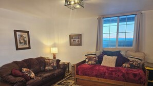 Una habitación acogedora con un sofá de cuero marrón, una cama de madera con una colcha roja y un ventanal con vistas al cielo y las montañas.