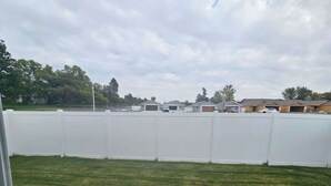 Une cour arrière clôturée avec une pelouse verdoyante, une terrasse en béton et une rangée de maisons en arrière-plan.