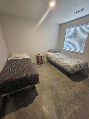 A small, single-room apartment with two beds, a small table, and a window with blinds.