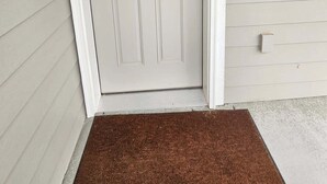 Une porte blanche avec une petite fenêtre, un paillasson et un interrupteur sur le mur.