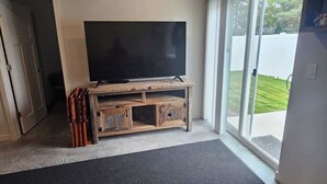 A living room with a flat-screen TV on a wooden TV stand, a black rug, and a sliding glass door leading to a backyard. - Cozy 2BR Home Parking and Full Kitchen (Stanfield)