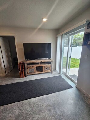 A living room with a flat-screen TV on a wooden TV stand, a black rug, and a sliding glass door leading to a backyard. - Cozy 2BR Home Parking and Full Kitchen (Stanfield)