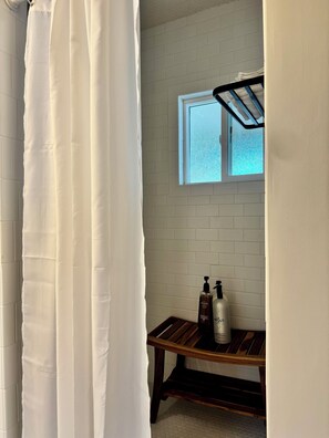 Un bagno con pareti rivestite di piastrelle bianche, una finestra, una panca di legno e due flaconi di prodotti per la cura dei capelli.