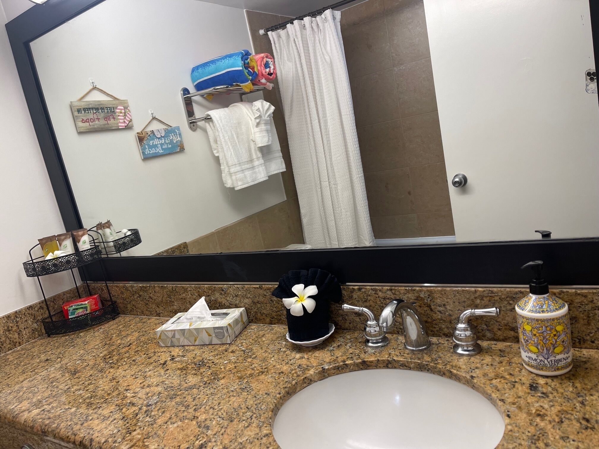 A bathroom with a granite countertop, a white sink, a mirror, a black towel, a tissue box, a hand soap dispenser, and a shower curtain.