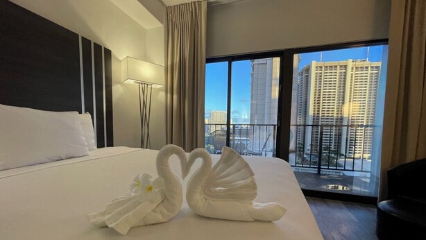 Habitación de hotel con una cama grande, vistas a edificios altos y un balcón con sillas.