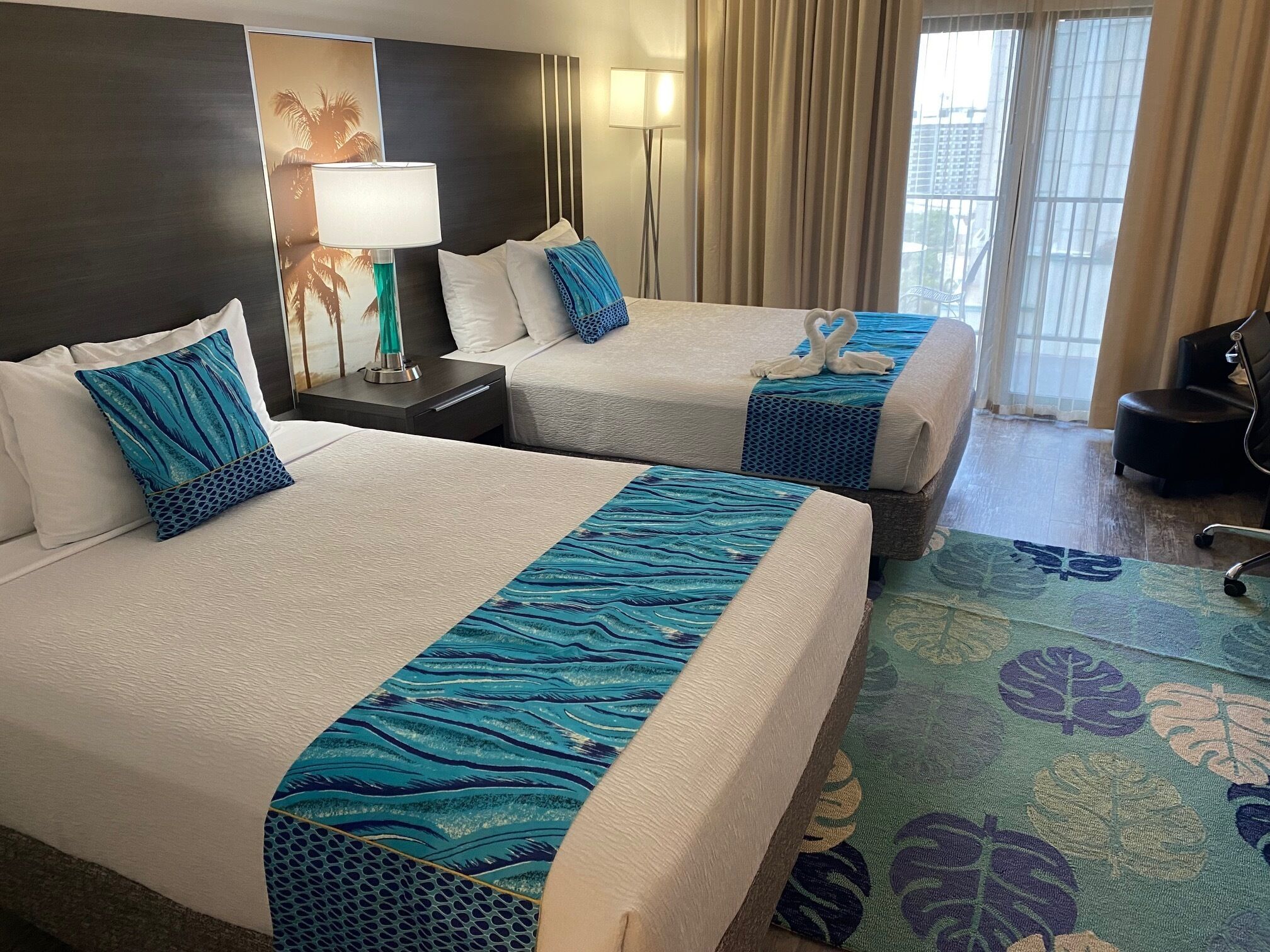Two hotel beds with matching blue and white bedding, a nightstand with a lamp, and a view of the city through large windows.