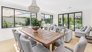 A spacious living area with a wooden dining table, grey upholstered sofas, and a large window offering a view of the outdoors. - Whitepepper Cottage, Pokolbin, Hunter Valley (Pokolbin)
